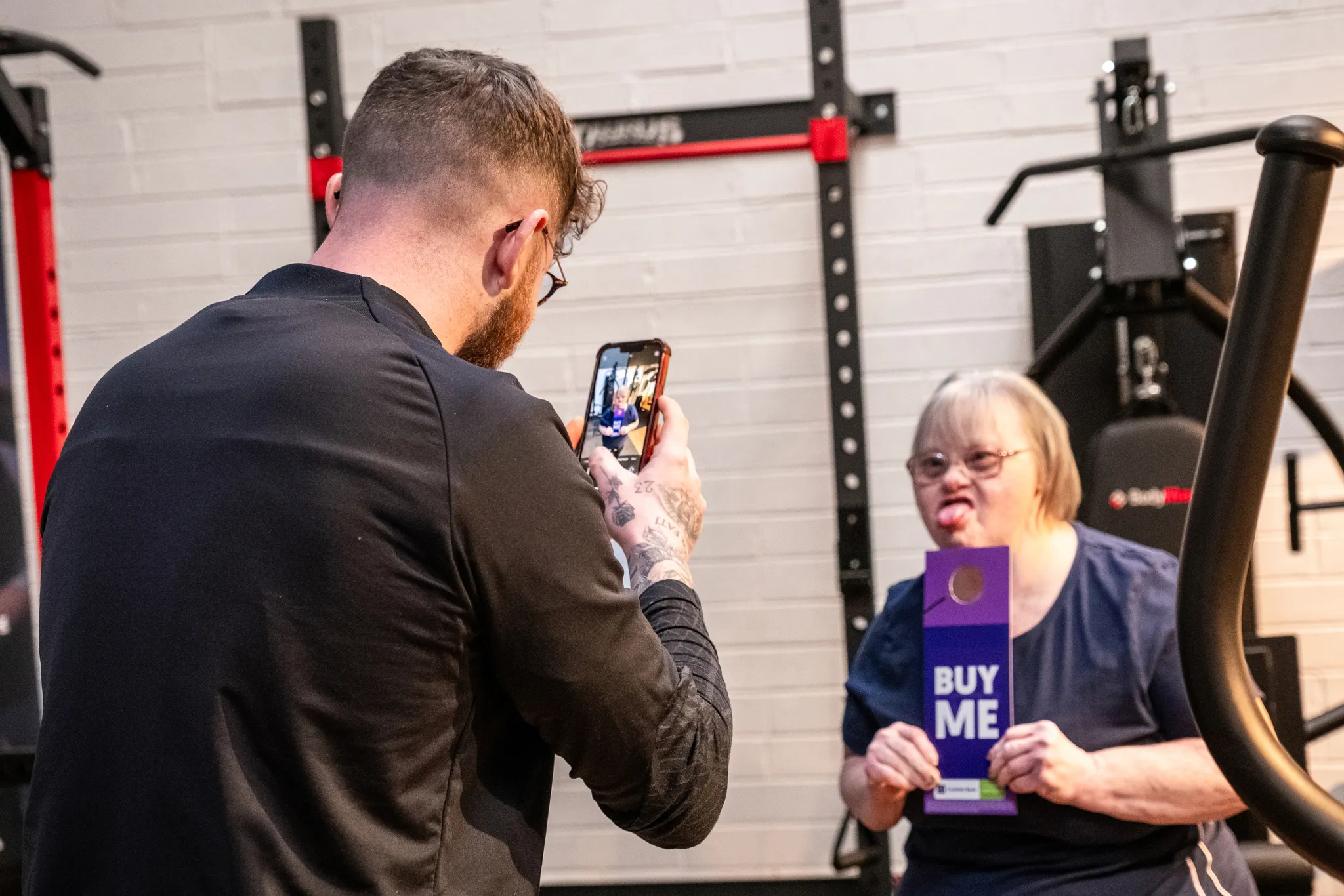 A tattooed man photographing a woman with Down's syndrome on a smartphone in a gym setting, as she playfully poses holding a purple Buy Me promotional card in front of gym equipment